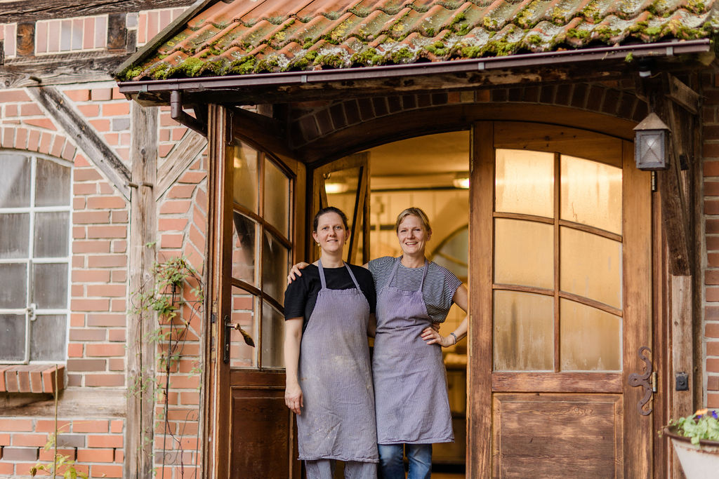 Zwei Bäckerinnen lächeln im Eingang der Backstube zwischen Fachwerk und Backsteinmauer
